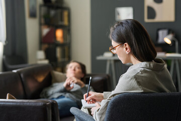 Counselor writing notes during therapy session with patient lying on couch in relaxing office environment, focusing on mental health and emotional well-being