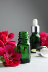 Bottles of geranium essential oil and beautiful flowers on white wooden table, closeup