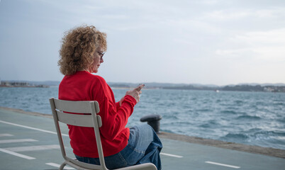 Fototapeta premium Woman in a red sweater sitting on a chair by the sea, using a smartphone