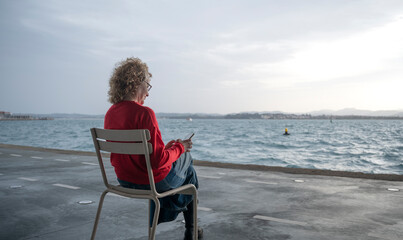 Fototapeta premium Woman sitting on a chair by the sea, using a smartphone, with a distant shoreline in view
