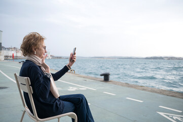 Fototapeta premium Elderly woman video calling by the seaside on a cloudy day