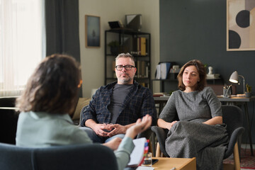 Couple sitting in office discussing relationship issues with therapist holding clipboard. Room has...