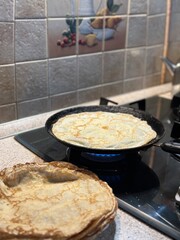 A cozy kitchen photo with a focus on making crepes. A stovetop with a black frying pan containing a cooking crepe, positioned on a lit gas burner. In the foreground, there is a stack of cooked crepes.