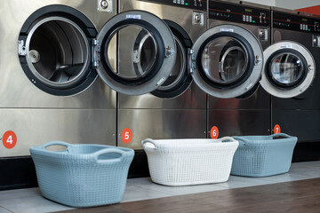 Empty washing machines built in row in self-service collective automatic laundry with baskets near...