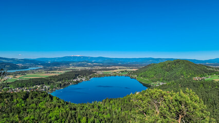 Breathtaking aerial view of a tranquil lake Klopein nestled amidst lush green forest in Carinthia, Austria. Crystal-clear water reflects vibrant blue sky. Rolling hills and mountains in Austrian Alps