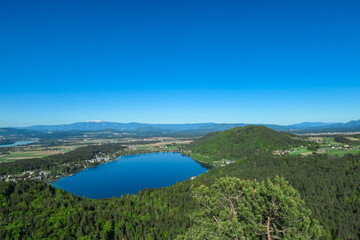 Breathtaking aerial view of a tranquil lake Klopein nestled amidst lush green forest in Carinthia, Austria. Crystal-clear water reflects vibrant blue sky. Rolling hills and mountains in Austrian Alps