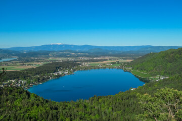 Breathtaking aerial view of a tranquil lake Klopein nestled amidst lush green forest in Carinthia, Austria. Crystal-clear water reflects vibrant blue sky. Rolling hills and mountains in Austrian Alps