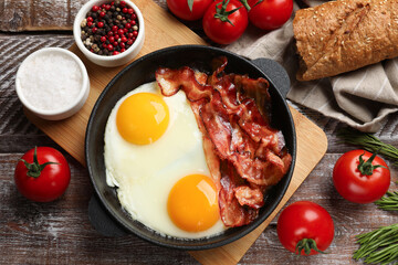 Tasty bacon and eggs in dish among products on wooden table, flat lay