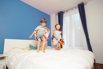 A pair of young kids with capes cheerfully bouncing on bed