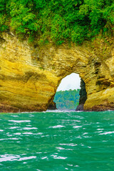 Natural rock arch formation along the Pacific coast, with lush green vegetation atop and turquoise water below, leading to a view of the forested landscape beyond. High-quality photo. Uvita Puntarenas