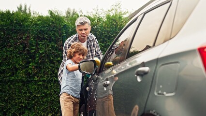 Adult man teaches pre-adolescent son right washing car at backyard on sunny day. Father and...