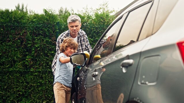 Adult man teaches pre-adolescent son right washing car at backyard on sunny day. Father and pre-teenage boy are using sponges and cleaning auto outdoor near home. Happy family concept.