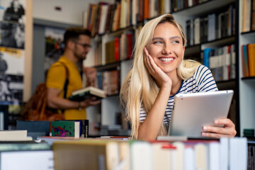 A radiant Caucasian young woman engages with a digital tablet in a library while a focused male student examines books in the background, surrounded by diverse literature.
