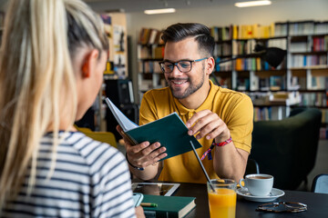 A cheerful Caucasian male and female engage in a lively discussion behind a library table, surrounded by an array of books, with refreshing beverages at their side.