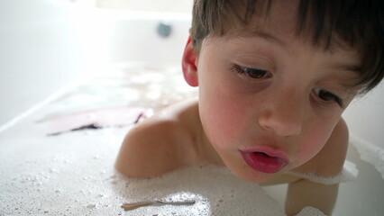Young boy resting in a bubble bath, blowing bubbles in the water. His relaxed posture and focused expression capture the joy and tranquility of childhood bath time moments
