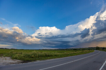 Fototapeta premium A scenic road cuts through a grassy field under a dramatic sky with cumulus clouds. The setting sun reflects warm tones of orange and pink, creating a stunning view