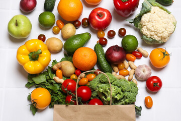 Vegan food delivery. Paper bag with different fresh vegetables, nuts and fruits on white tiled table, flat lay