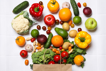 Vegan food delivery. Paper bag with different fresh vegetables, nuts and fruits on white tiled table, flat lay