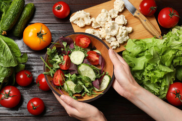 Healthy vegetarian food. Woman holding bowl of salad at wooden table with vegetables, top view