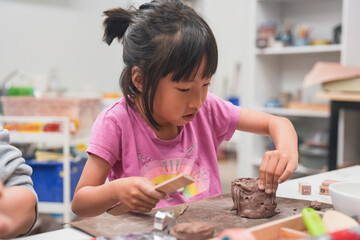 Young girl intensely focuses on shaping a piece of clay with wooden tool during art class. Her dedication to the creative process emphasizes the importance of hands-on learning and artistic expression