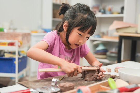 Young girl intensely focuses on shaping a piece of clay with wooden tool during art class. Her dedication to the creative process emphasizes the importance of hands-on learning and artistic expression
