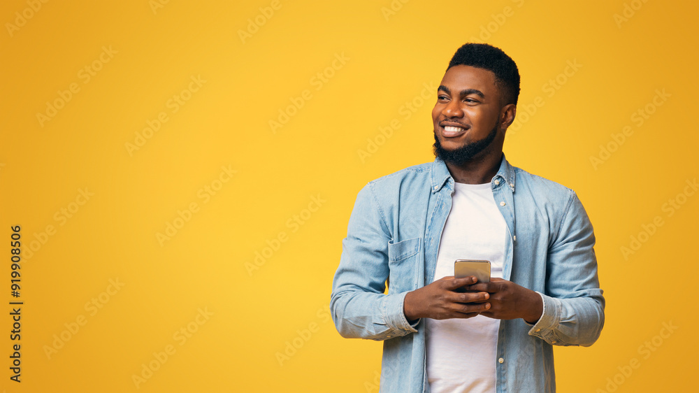 Poster handsome black man holding smartphone and looking aside at copy space on yellow studio background