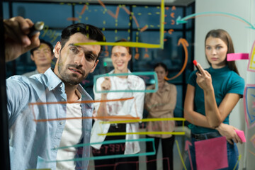 Professional businessman writing on glass wall at night office while coworker listening carefully. Group of business team working together,brainstorming, sharing marketing idea. Business plan.Tracery.