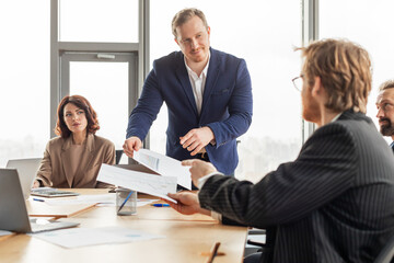 A businessman stands at the head of a conference table, distributing papers to a colleague seated at the table. Other colleagues sit at the table, listening to the businessman.