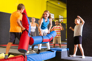Focused smiling teenage girl fighting by soft pugil sticks with boy while standing on inflatable...