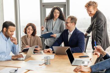 A group of business professionals, including men and women, work together at a large wooden table in a modern office. They are looking at laptops, taking notes, and discussing ideas.