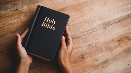 Close-up of a woman's hands gently holding a closed book with the text "Holy Bible" on a wooden table. The image reflects a moment of spiritual reflection, emphasizing the importan