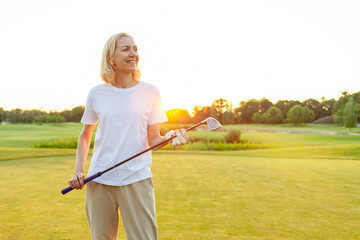 An elderly woman with a smile on her face is standing on a golf course holding a golf club. He is...