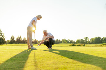 elderly couple seniors playing golf on golf course at sunset, old man and woman practicing with golf clubs in nature, husband and wife in white uniform actively relaxing