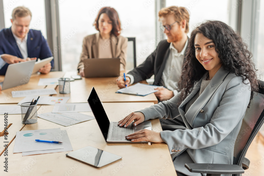 Poster a woman with curly hair sits at a conference table and types on her laptop while other business prof