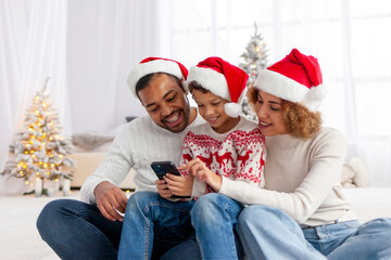 african american family in santa hat hugging at home for christmas and giving smartphone to son, teenage boy celebrating new year with mom and dad and using phone, parents choosing online and typing