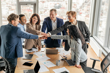 A diverse team of business professionals stand around a conference table, their hands stacked in...