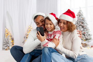 african american family in santa hat hugging at home for christmas and giving smartphone to son, teenage boy celebrating new year with mom and dad and using phone, parents choosing online and typing