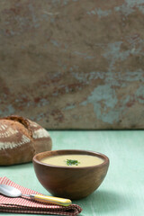 A yellow vegetable soup piping hot in a carved wooden bowl on a green vintage table with red checker napkin and spoon and rustic sour dough  loaf of bread in the background