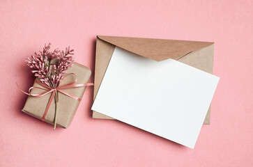 Blank greeting card mockup, white paper card with envelope, gift box and flowers on pink background