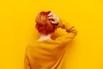 puzzled red-haired teenage boy thinks and chooses on a yellow isolated background, confused child with an orange hairstyle remembers and scratches the back of his head, rear view