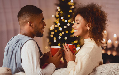 Excited black couple drinking coffee on Christmas eve, enjoying cozy evening at home