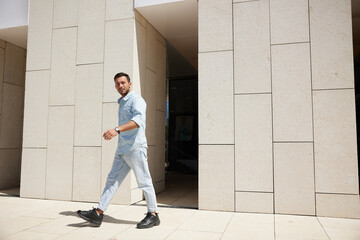 Confident young man walking outdoors in a modern urban setting, wearing casual light-colored denim, exuding confidence and style