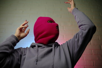 Individual in a pink balaclava poses against a colorful backdrop, making a gesture