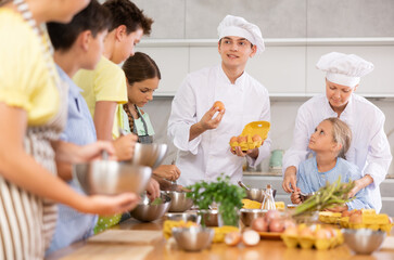 Young guy and adult woman cook in uniform teaches group of children how to cook dish