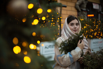 Street city photo. Day. Young adult child free woman in woolen scarf on her head at winter holiday fair holds green fir branch in hand. Christmas tree. Yellow bokeh lights. Happy New Year 2025. Joy