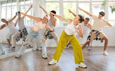 Young girl in bright clothes, together with group of teenage friends, learn to dance hip hop in choreographic hall. Students move to modern music, do moonwalking, squats, bends, work with body