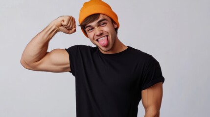 Naklejka premium Happy gen Z young man wearing black t-shirt and orange beanie flexing his bicep and sticking out tongue. Studio shot, white background