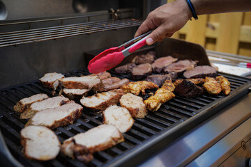 A person uses tongs to grill a variety of meats on an outdoor barbecue grill. The meats include chicken, steak, and pork, arranged appealingly on the grill.