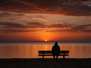 Obraz premium A dark silhouette of a person sitting alone on a bench, facing the horizon during a sunset with vibrant orange and red skies.