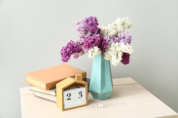 Vase of beautiful lilac flowers with calendar and books on table near white wall